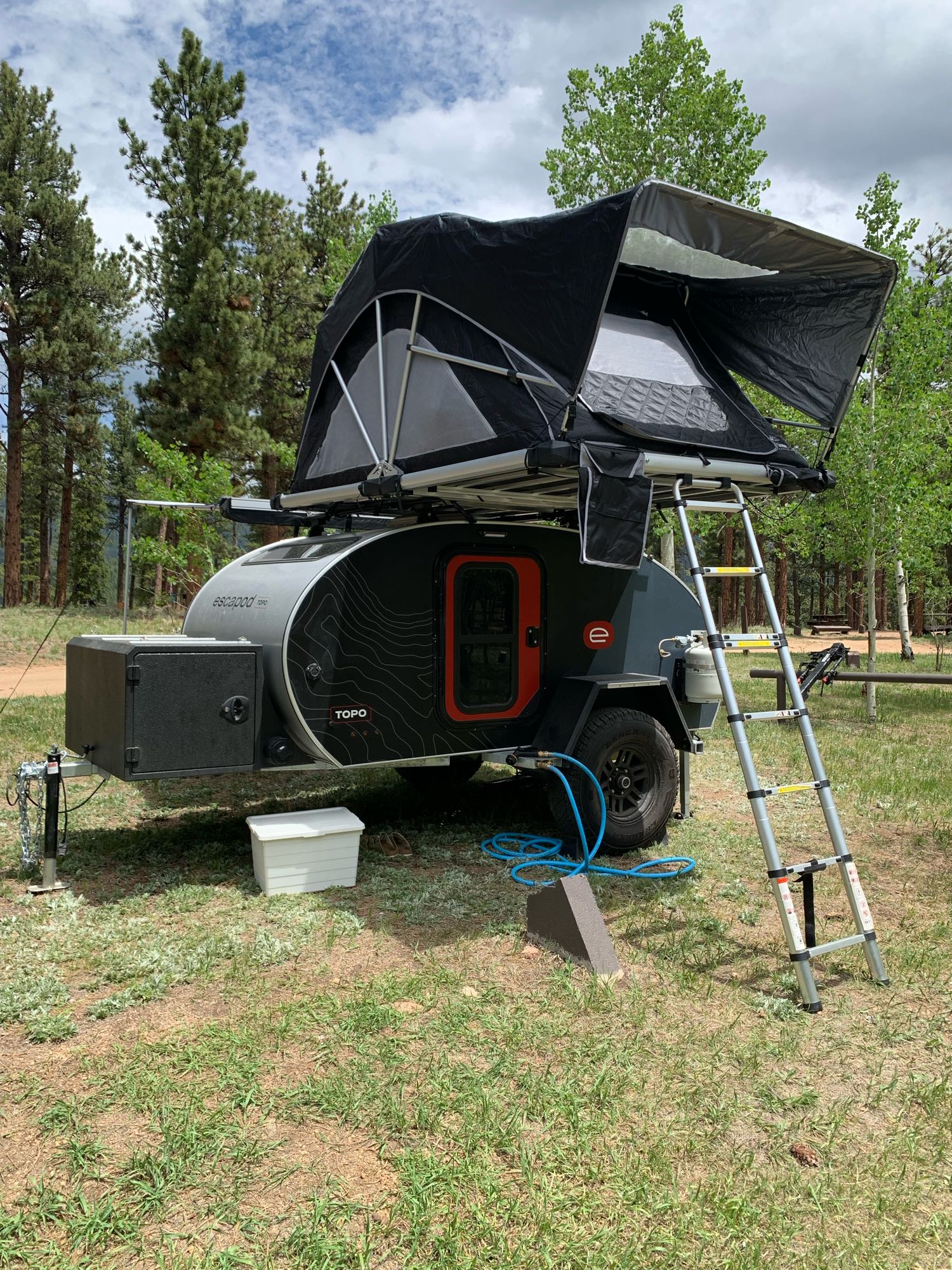 An Escapod topo teardrop trailer with a rooftop tent set up for an rv rental camping adventure in a forest.