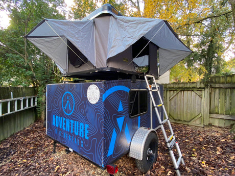 This RV rental features a dark blue trailer with a deployed rooftop tent and ladder, ideal for camping adventures.