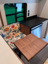 Cozy RV rental interior with floral dinette, fold-out table, compact fridge, and window view. Perfect for your next adventure!
