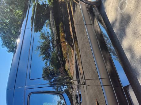 Shiny black RV rental side reflecting blue sky, green trees, and pavement. A sun glare adds sparkle.