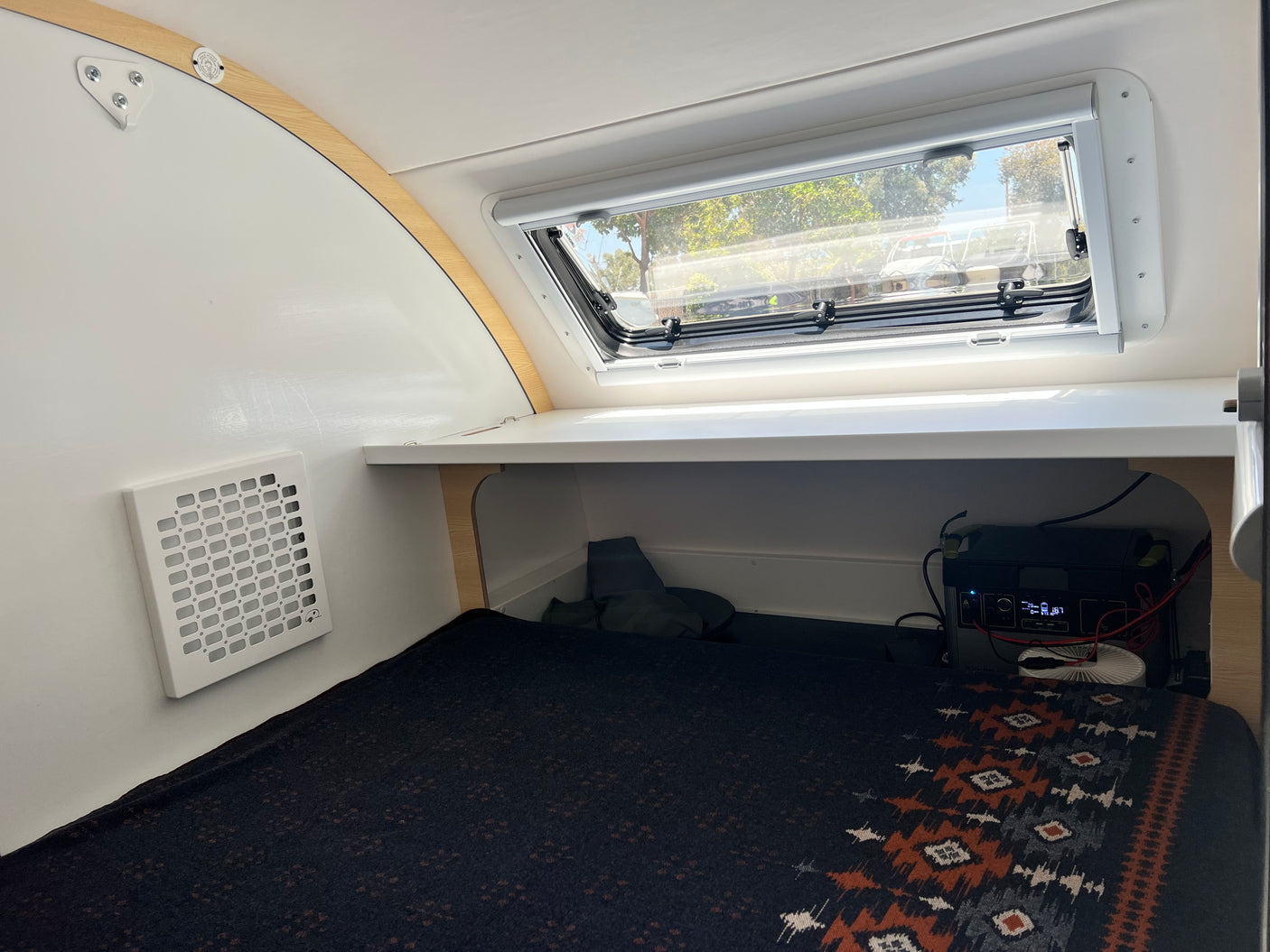 Small RV rental interior: cozy bed with patterned blanket, large window, and portable power station.