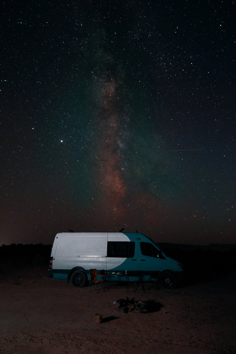 An RV rental camper van parked at night under a brilliant Milky Way and stars. Camp chairs and fire pit visible.
