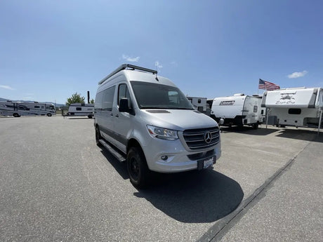 A silver Mercedes-Benz Sprinter van, an ideal RV rental, parked in a sunny outdoor lot with other RVs under a clear sky.