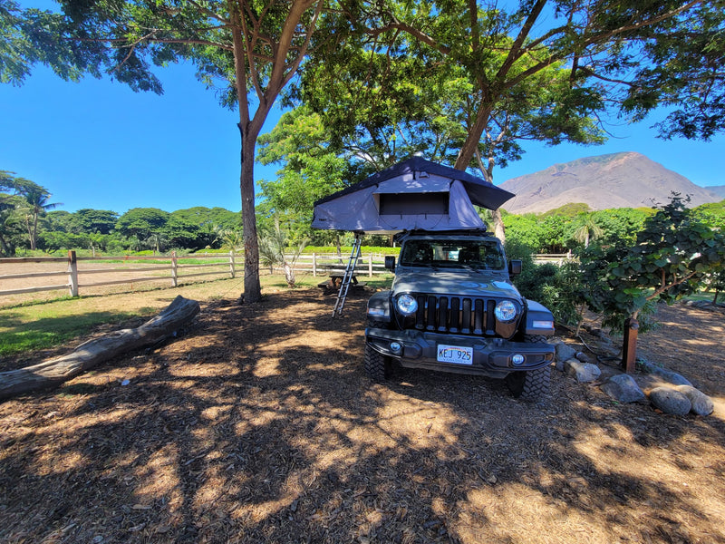 Green Jeep Wrangler with rooftop tent (an RV rental setup) under shade trees, facing a mountain with a fenced field nearby. — photo 4
