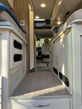 Spacious RV rental interior featuring light wood cabinetry, a sleeping area, and a clear view to the driver's cabin and outdoors.