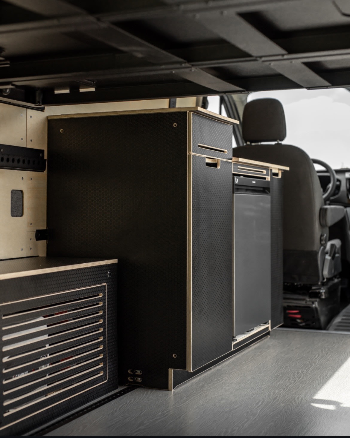 Compact rv rental van interior with modern black textured modular cabinets, integrated fridge, and light wood trim details.