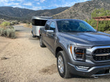 A gray Ford F-150 truck tows a silver Airstream Basecamp RV on a dirt road in the mountains. Explore with an rv rental!