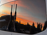 Golden sunset and mountain silhouettes reflected on an RV rental's rear window, framed by wipers. Experience serene travel views.