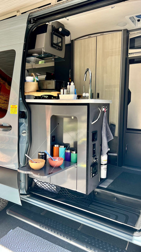 Compact and functional RV rental kitchen in a camper van, featuring a sink, storage, and pull-out counter for mobile living.