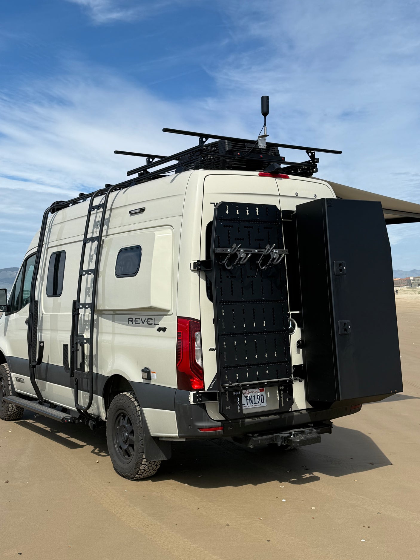 White Winnebago Revel 4x4 rv rental camper van on a sandy beach, showcasing its roof rack, side ladder, and rear storage system.