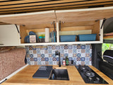 RV rental: A modern camper van kitchen with patterned tile backsplash, wooden counter, sink, stove, and open cabinets.