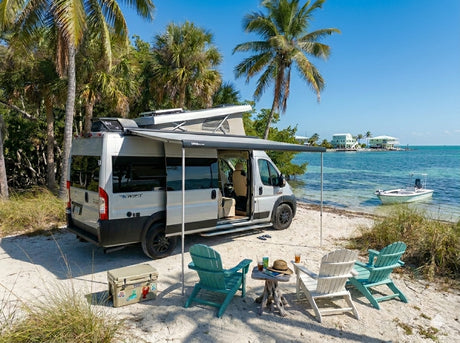 Enjoy the perfect beach getaway in an RV rental! Camper van parked on a sandy shore with chairs, an awning, and ocean views.