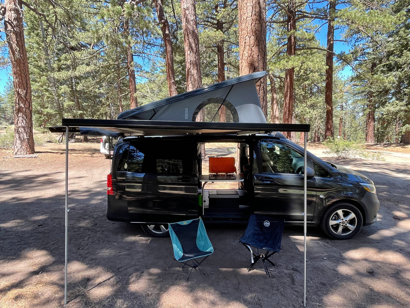 RV rental camper van with extended pop-top and awning, featuring a kitchen setup and chairs, parked in a sunny forest.