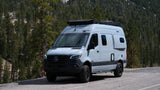 Adventure-ready Mercedes-Benz Sprinter camper van RV rental parked on a mountain road with a dense forest backdrop.