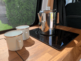 Morning coffee in an rv rental: Two enamel mugs and a percolator on an induction cooktop, with a screened window view of trees.