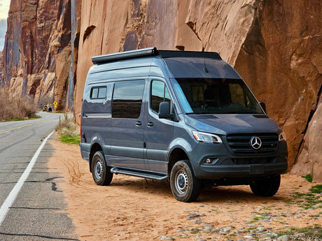 Dark grey Mercedes-Benz Sprinter camper van (RV rental) parked on a dirt road shoulder beside towering red rock cliffs.