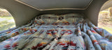 Inside an RV rental: a light blue, fluffy patterned blanket on a cozy pop-top bed, with outdoor views.