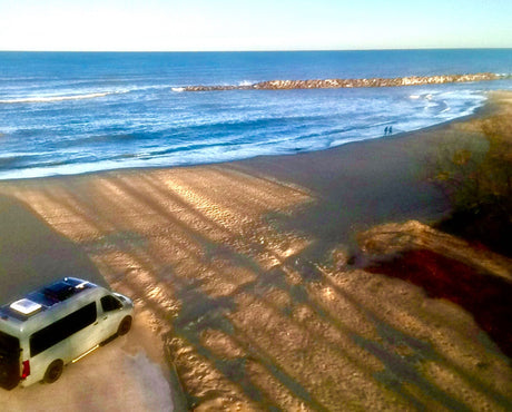 RV rental parked on a sunny beach with striking long shadows. The ocean and a jetty are in the background, with two people walking.