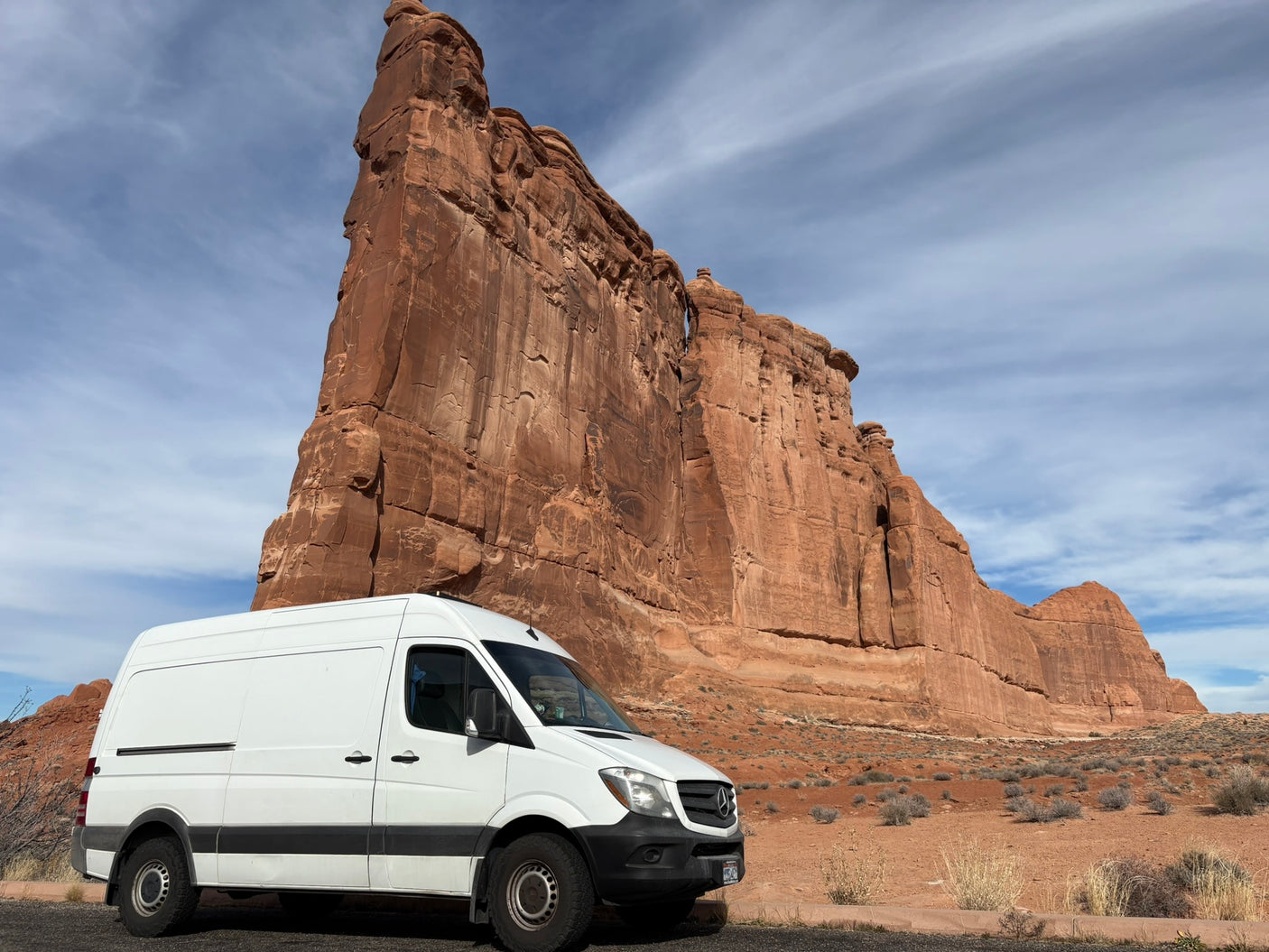 White Sprinter rv rental parked by a colossal red rock formation in a national park. Ideal for desert road trips.