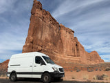 White Sprinter rv rental parked by a colossal red rock formation in a national park. Ideal for desert road trips.