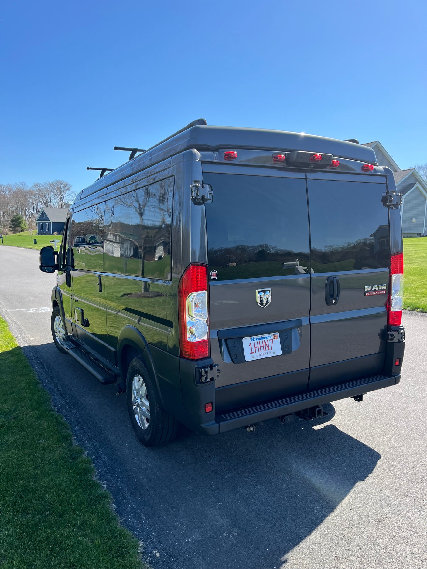 Dark gray Ram ProMaster camper van, perfect for RV rental, with roof rack and rear hitch, parked on a sunny road.