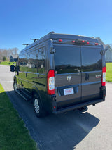 Dark gray Ram ProMaster camper van, perfect for RV rental, with roof rack and rear hitch, parked on a sunny road.