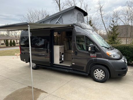 Modern dark grey pop-top rv rental with extended awning and open door revealing interior kitchen, parked on a driveway.