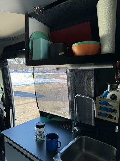 RV rental kitchen: compact sink, coffee, and storage inside a van, with a snowy outdoor view.