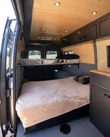 Spacious RV rental interior featuring comfortable bunk beds, sleek overhead cabinets, and a compact kitchen area in a modern camper van.