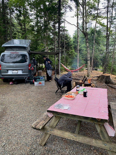 A gray Mercedes-Benz rv rental camper van at a peaceful campsite with a fire pit, red hammock, and picnic table set for a meal.
