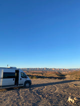 RV rental camper van parked on a dirt road overlooking vast desert mountains. Clear sky, small dog exploring scenic views.