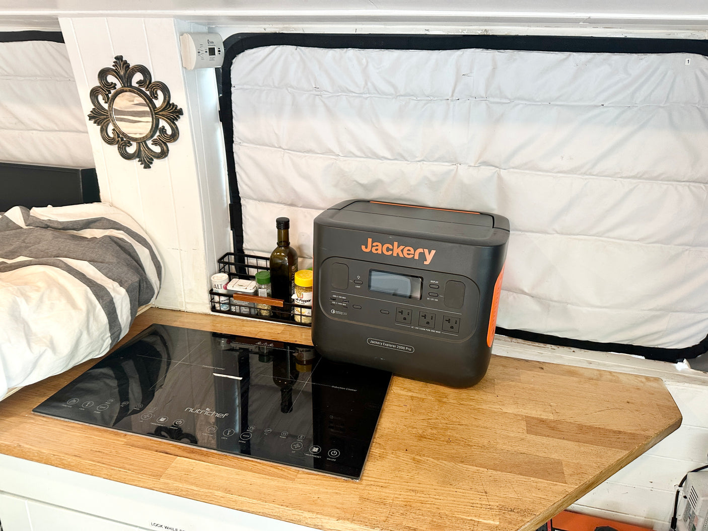 Compact RV rental interior featuring a Jackery power station, Nutrichef induction cooker, and cozy bed setup.
