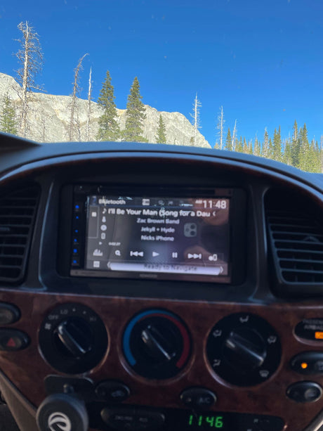 RV rental dash with Zac Brown Band playing. Stunning mountain landscape, dead and evergreen trees under a clear blue sky, ideal road trip.