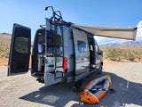 Your RV rental adventure awaits! Grey camper van with open doors, deployed awning, and kayak in a scenic desert.