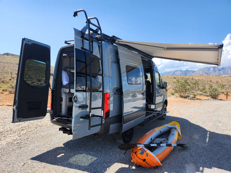 Your RV rental adventure awaits! Grey camper van with open doors, deployed awning, and kayak in a scenic desert.