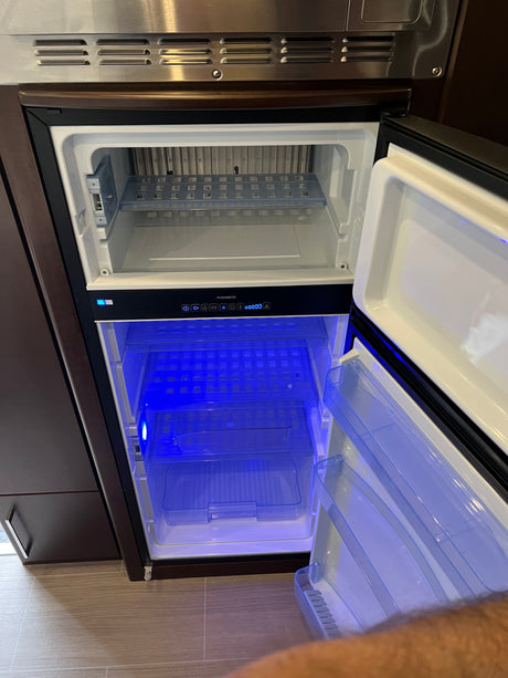 Open Dometic refrigerator and freezer in an RV rental, showing blue interior lights and empty shelves, ready for travel storage.