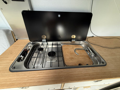 RV rental kitchenette: two-burner stove, sink with faucet, and wood cutting board cover on a bamboo counter in a camper van.