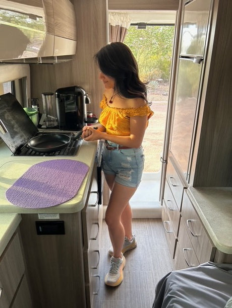 A woman in a yellow top cooks in a compact rv rental kitchen, stirring a pan on the stove. Ideal for road trip adventures.