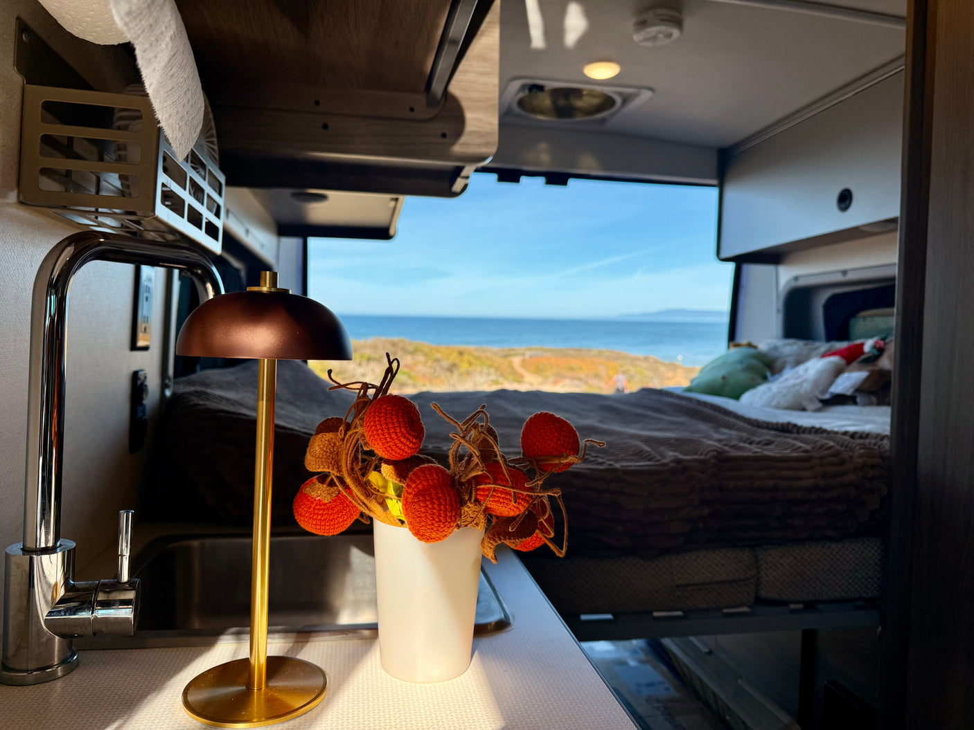 An RV rental offers a stunning ocean view from a cozy bed, complemented by a compact kitchenette and autumn decor.