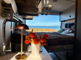 An RV rental offers a stunning ocean view from a cozy bed, complemented by a compact kitchenette and autumn decor.