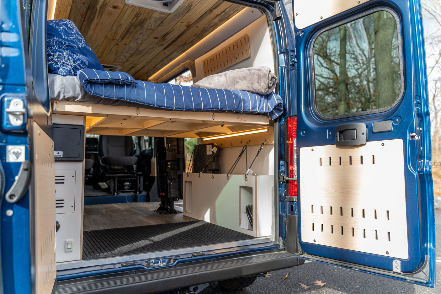 Inside a blue RV rental with open rear doors. Features a raised bed with blue bedding, custom wood ceiling, and lower storage.