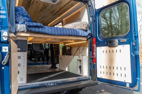 Inside a blue RV rental with open rear doors. Features a raised bed with blue bedding, custom wood ceiling, and lower storage.