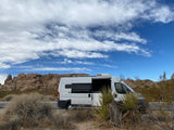 White RV rental parked in a desert landscape with its side door open, surrounded by large rocks and a blue sky.