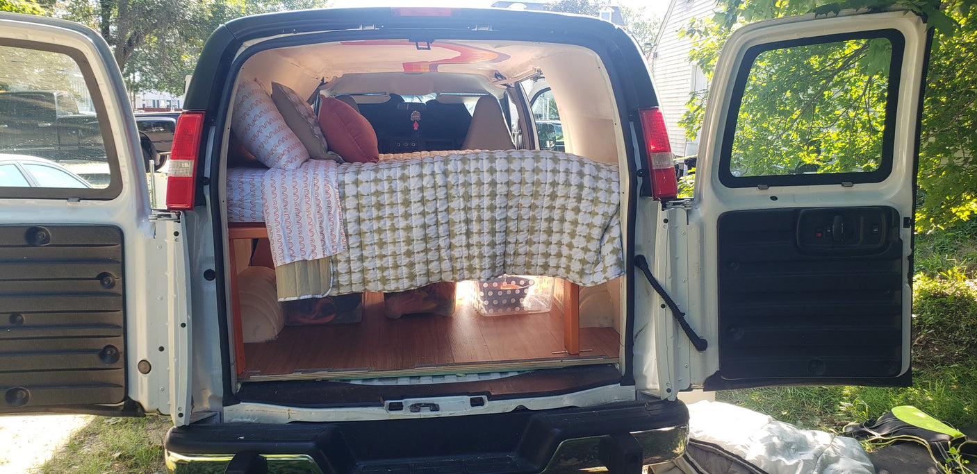 Converted van RV rental interior: cozy bed with pillows, patterned bedding, & under-bed storage, with open rear doors.