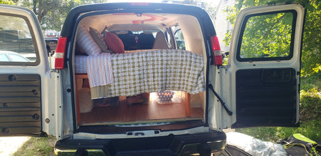 Converted van RV rental interior: cozy bed with pillows, patterned bedding, & under-bed storage, with open rear doors.