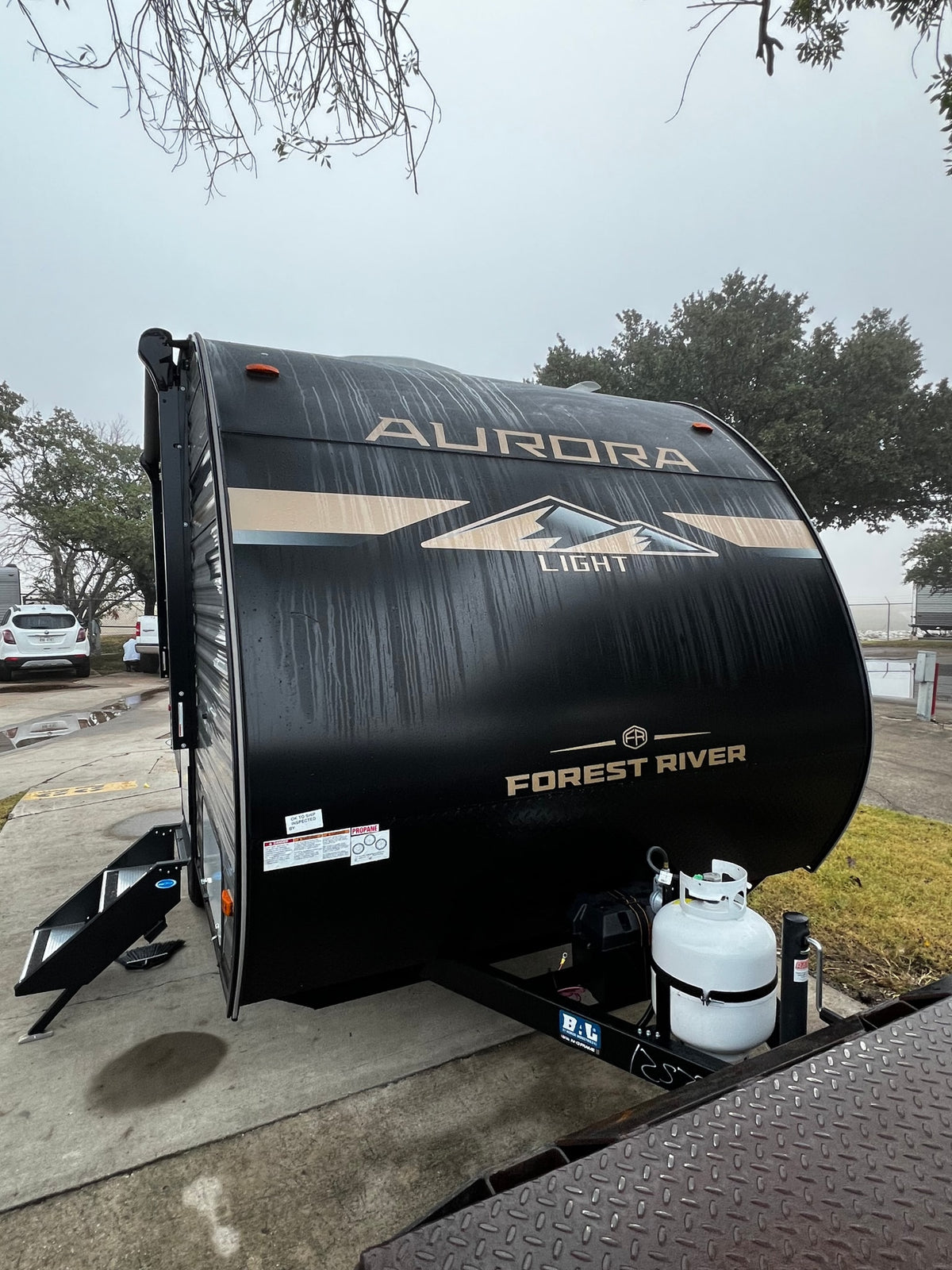 A black Forest River Aurora Light rv rental travel trailer, with propane tanks, is parked on concrete under a cloudy sky, ready for camping. — photo 11