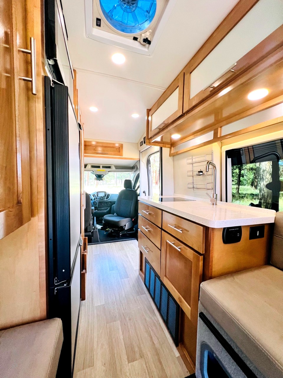 This modern rv rental interior features a compact kitchen with wood cabinets, sink, fridge, and seating, leading to the front cabin. — photo 9