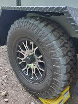 Close-up of an RV rental trailer's off-road tire with a custom wheel, resting on a yellow leveling block. Diamond plate fender above.