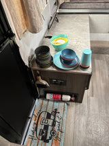 RV rental kitchen: camping dinnerware, pots, cups, and mixing bowls on the counter. Fire extinguisher and "Home is where you park it" mat.