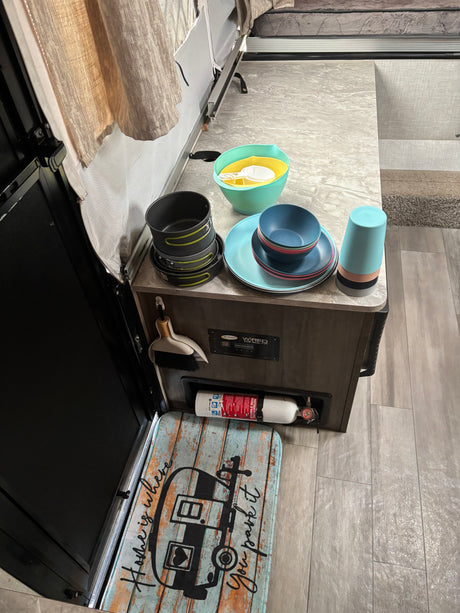 RV rental kitchen: camping dinnerware, pots, cups, and mixing bowls on the counter. Fire extinguisher and "Home is where you park it" mat.